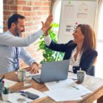 An Auckland insurance broker and client high-fiving across a desk after a successful advice meeting, laptop and paperwork visible