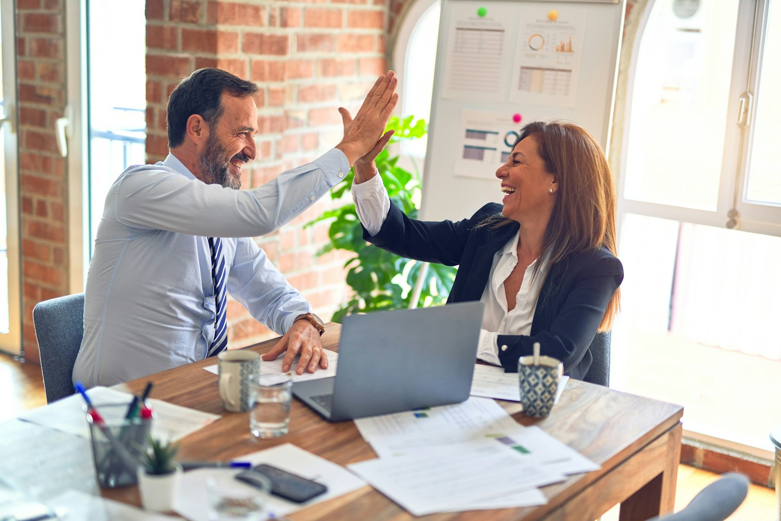 An Auckland insurance broker and client high-fiving across a desk after a successful advice meeting, laptop and paperwork visible